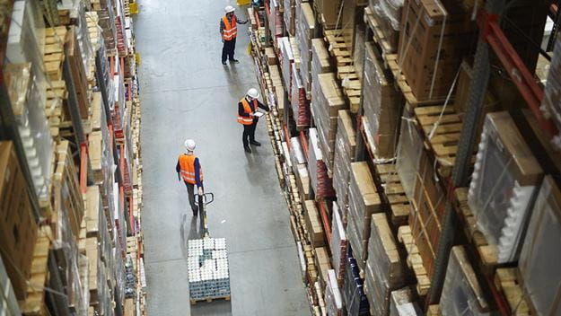 Above view of warehouse workers moving goods and counting stock in aisle between rows of tall shelves full of packed boxes.