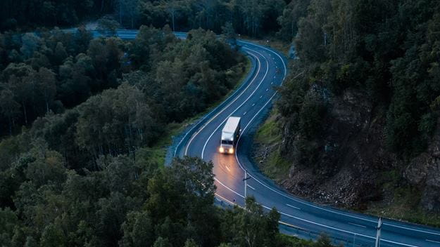 Un camión circula por una carretera sinuosa rodeada de un denso bosque al anochecer.