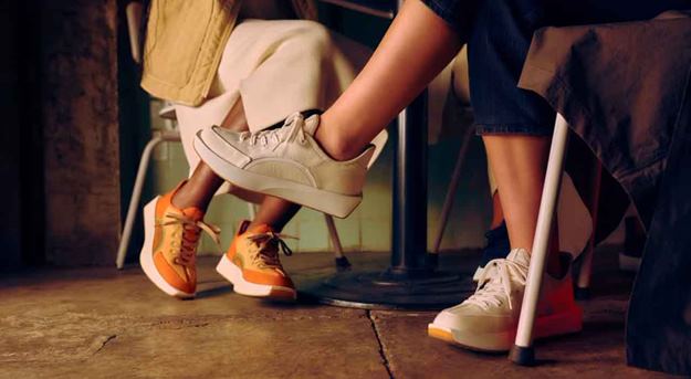 Two pairs of legs seen from the knees down, wearing casual sneakers. One person is wearing beige sneakers with white soles, while the other is in bright orange and white sneakers. They are seated at a small table in a dimly lit indoor setting with a concrete floor.