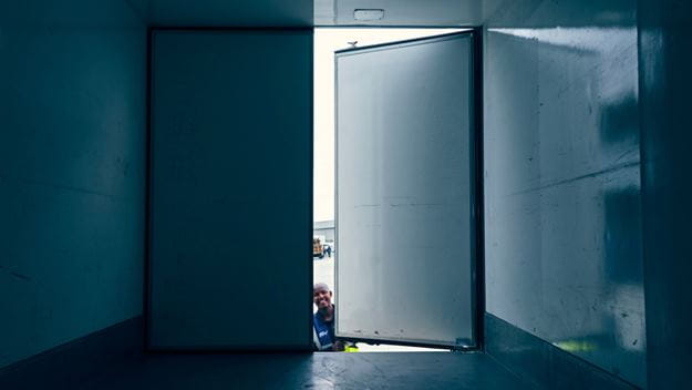 Een open deur in een grote, lege container met minimale verlichting, die een glimp van de buitenwereld laat zien