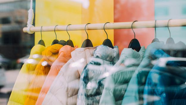 A row of colourful puffer jackets hanging on black hangers, displayed on a wooden rack in a storefront window.