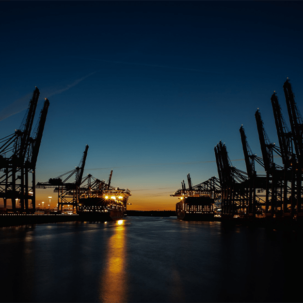 View of a port at dusk, with large cargo cranes silhouetted against a fading orange and blue sky