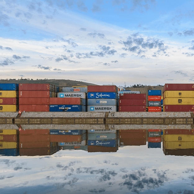 A row of colourful shipping containers stacked along a waterfront, reflecting in the calm water under a partly cloudy sky.