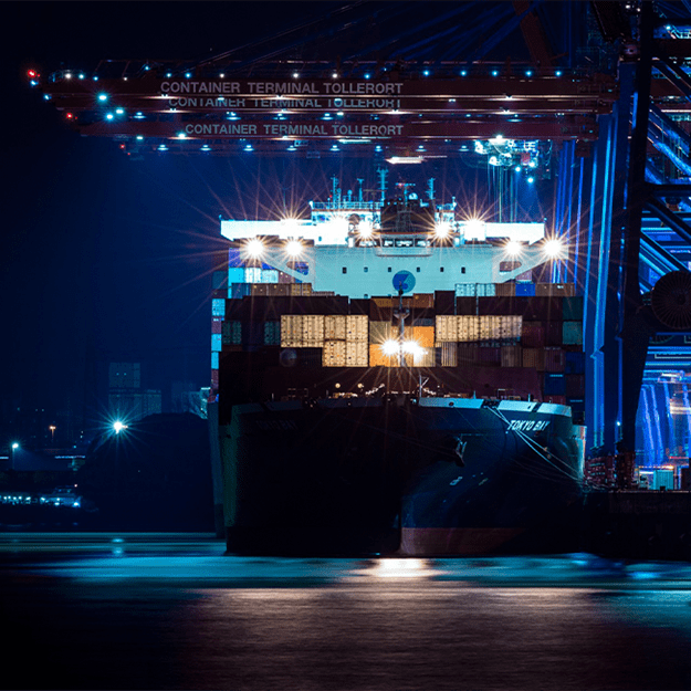 A large cargo ship docked at a container terminal at night, illuminated by bright blue and white lights, with port cranes towering above.